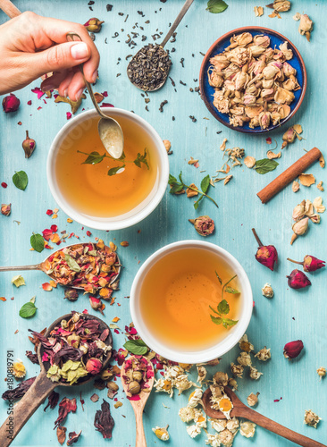 Two cups of healthy herbal tea with mint, cinnamon, dried rose and camomile flowers in spoons and man's hand holding spoon of honey, blue background, top view