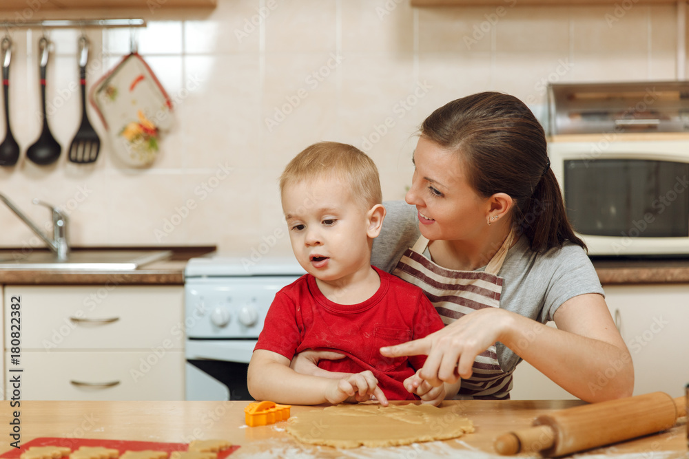 Fototapeta premium Little kid boy helps mother to cook Christmas ginger biscuit in light kitchen. Happy family mom 30-35 years and child 2-3 roll out dough and cut out cookies at home. Relationship and love concept