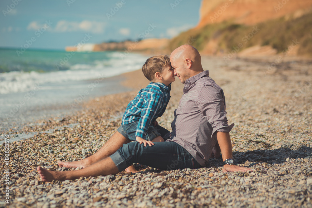 Touching appealing scene of father and son enjoy summer vacation together playing on stone beach wearing stylish shirt and fashion blue jeans both barefoot with adore landscape on background