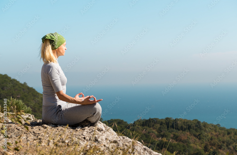 Beautiful blond yoga woman sitting on the top of the mountain in lotus pose. Meditation on the edge with a scenic view of the landscape and the sea.