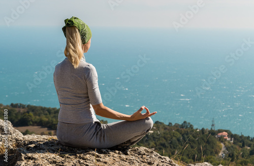 Beautiful blond yoga woman sitting on the top of the mountain in lotus pose. Meditation on the edge with a scenic view of the landscape and the sea.