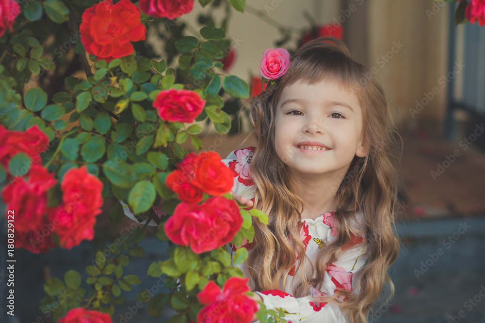 Cute baby girl with blond curly hairs and happy bright child eyes posing in central park close to huge bush of flowers red white pink roses wearing summer stylish clothes Cute baby girl with blond curly hairs and happy bright child eyes posing in central park close to huge bush of flowers red white pink roses wearing summer stylish clothes