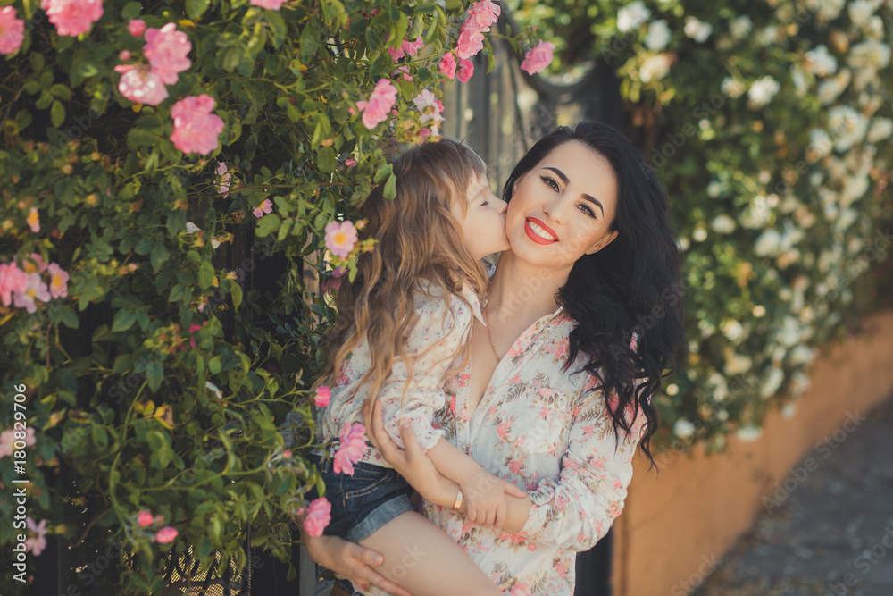 Best girlfriends adorable mother with dark black hairs and cute baby daughter with blond hairs and pink cheeks posing sit for together in central park close to huge bush of flowers roses Best girlfriends adorable mother with dark black hairs and cute baby daughter with blond hairs and pink cheeks posing sit for together in central park close to huge bush of flowers roses