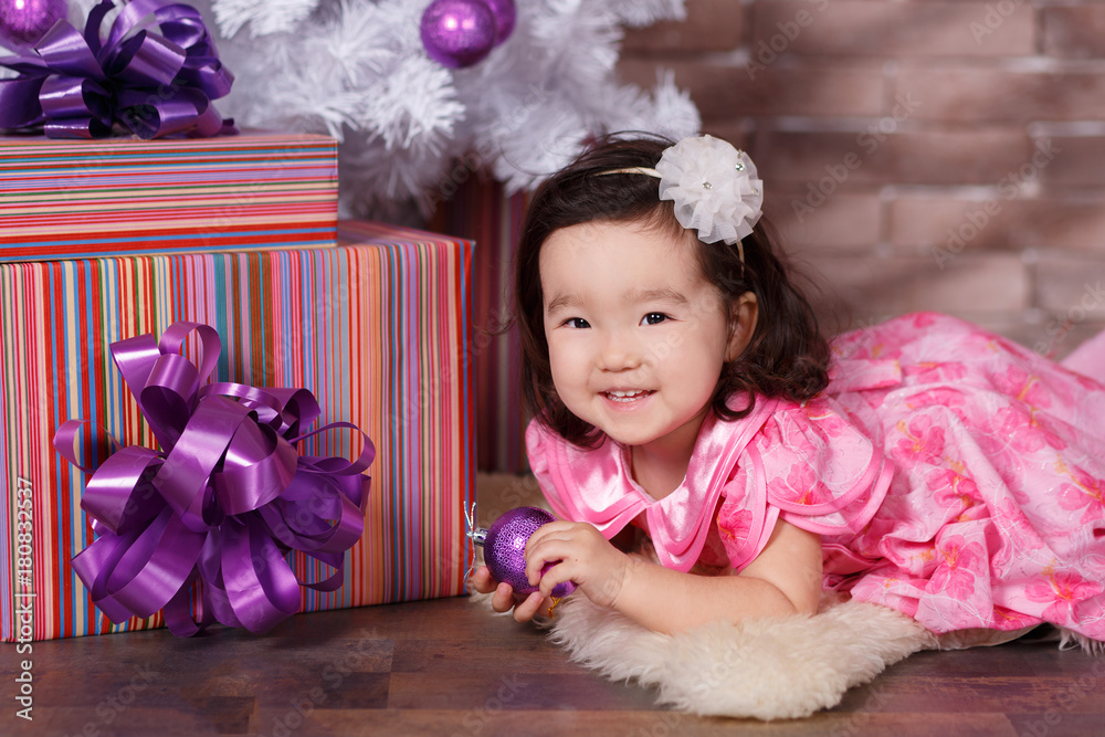 Asian pin up little cute girl wearing pink casual dress posing close to new  year christmas white tree with violet purple balls toys and colourful  presents Stock Photo
