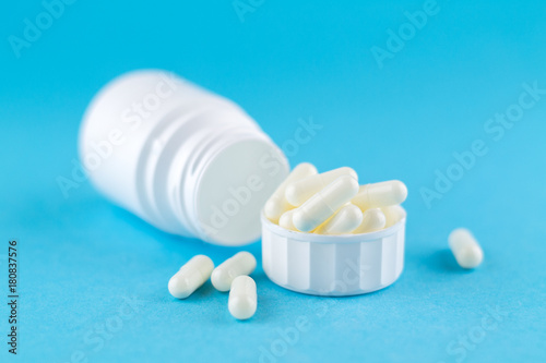 Close up white pill bottle with spilled out pills and capsules in cap on blue background with copy space. Focus on foreground, soft bokeh. Pharmacy drugstore concept