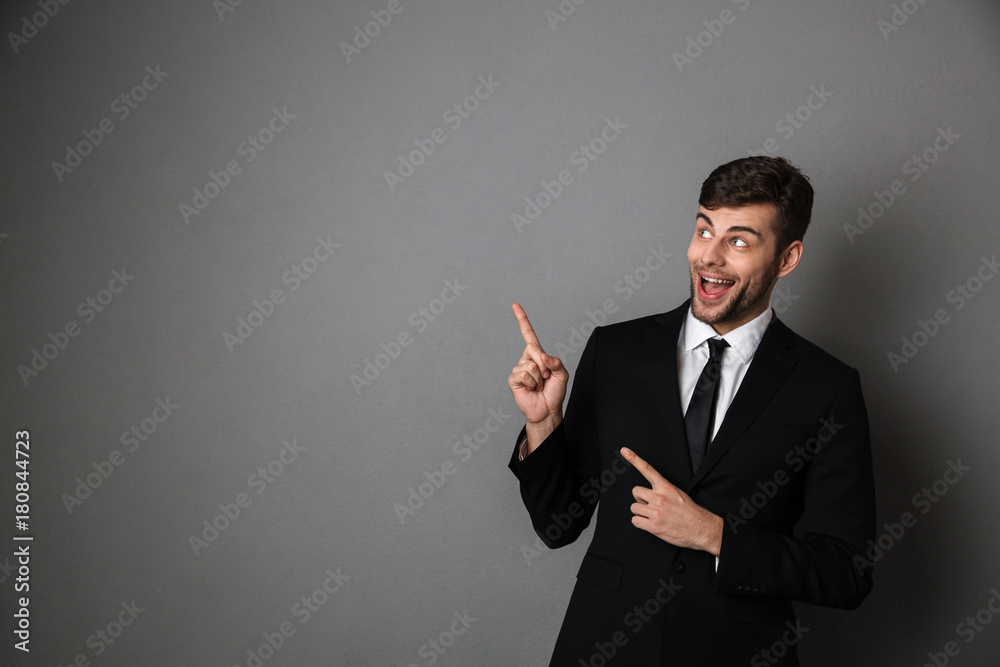 Overjoyed brunette man in classic suit pointing with two fingers, looking aside Overjoyed brunette man in classic suit pointing with two fingers, looking aside