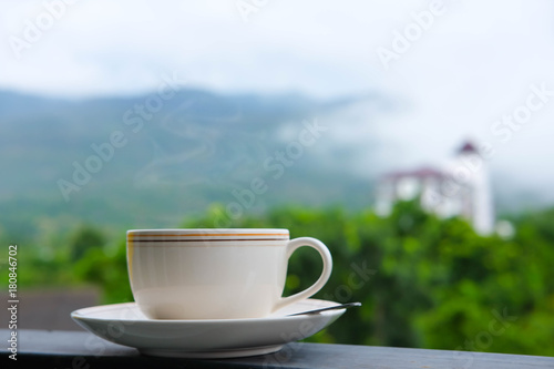 White cup of coffee on plate with smoke above on top and there are moantain and high building and forest in the background