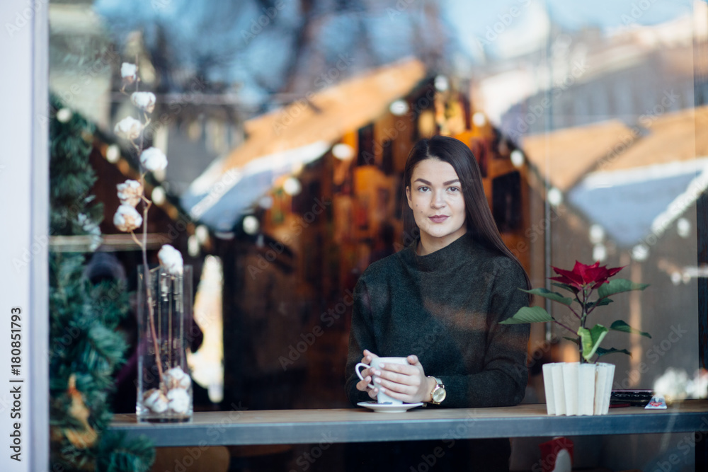 Beautiful brunette young woman in a cafe holding a cup of coffee or cocoa, seen through the window with buildings and lights reflections. She is looking away. Lifestyle concept.