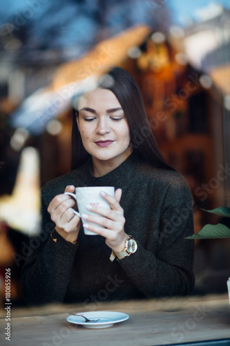 Beautiful brunette young woman in a cafe holding a cup of coffee or cocoa, seen through the window with buildings and lights reflections. She is looking away. Lifestyle concept.