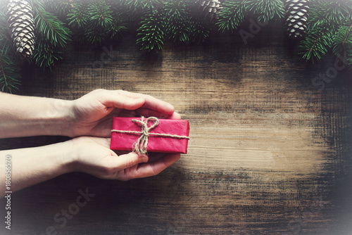 Red gift box in female hands and christmas fir tree on wooden background. Christmas celebration concept. © isavira