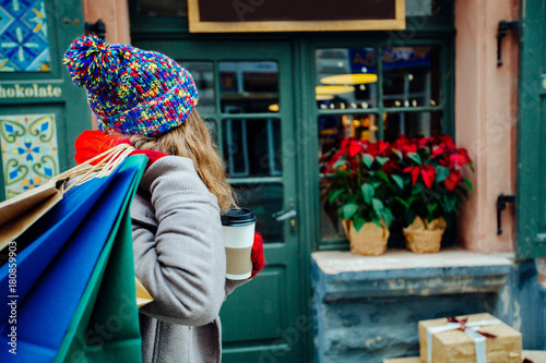 Attractive cute girl in with the shopping bags and holding disposable cup with warm drink in her hand. Female dressed in warm bright hat, red scarf and mittens. Happy holiday and freedom concept.