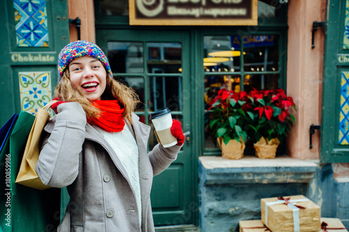 Attractive cute girl in with the shopping bags and holding disposable cup with warm drink in her hand. Female dressed in warm bright hat, red scarf and mittens. Happy holiday and freedom concept. © Iryna