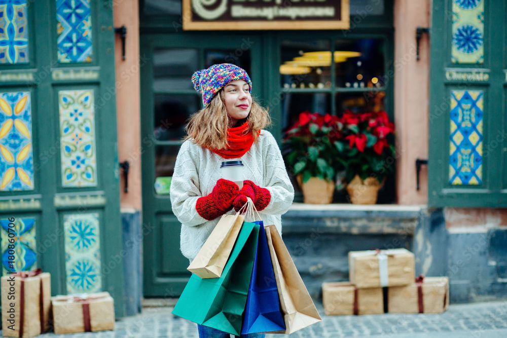Attractive girl with shopping bags holding disposable cup with warm drink in her hand.Woman near the shop with gifts enjoying winter and dressed in warm bright hat, red scarf and mittens.