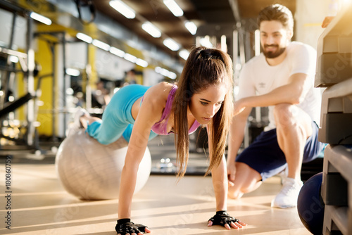 Attractive young athletic girl doing push ups with fitness ball in the modern gym next to the handsome satisfied personal trainer.