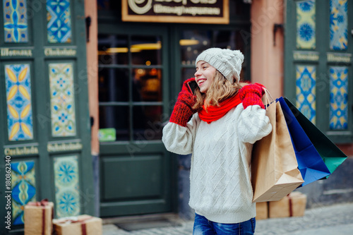 Beautiful smiling woman with shopping bags near shop with gift calling smart phone. Girl dressed in knitted hat, red scarf and mittens. Vacation, weekend, takeaway drinks, leisure concept © Iryna