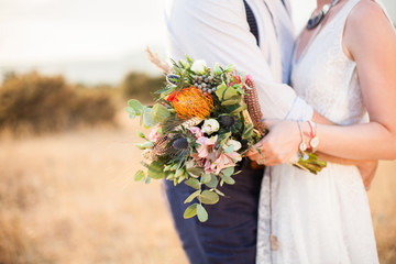 The bride and groom are hugging, holding wedding bouquet from orange, whire, pink flowers and standing on background of the autumn nature after wedding ceremony. Bride is dressed in white lace dress.