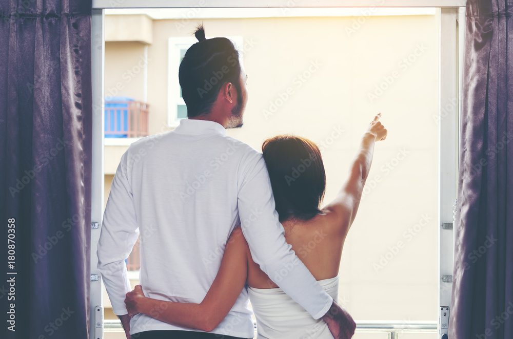 Obraz premium A married couple in white clothes standing near the window hugging on back side. Man with a beard and a woman are standing hugging each other. The girl and the boy are hugging.