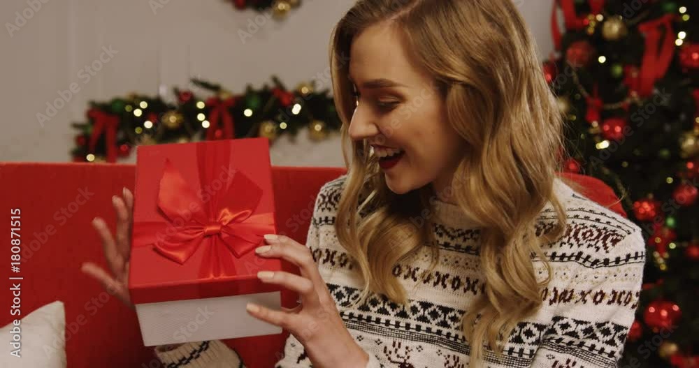 Attractive blonde young woman shaking her Christmas present and opening it with surprised eyes while sitting on the red sofa in the room on the Christmas tree background. Indoor. Close up. Portrait Attractive blonde young woman shaking her Christmas present and opening it with surprised eyes while sitting on the red sofa in the room on the Christmas tree background. Indoor. Close up. Portrait