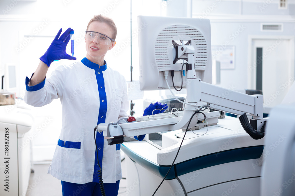 Transferring results. Attractive attentive female lab assistant standing near advanced medical apparatus while holding test tube and typing Transferring results. Attractive attentive female lab assistant standing near advanced medical apparatus while holding test tube and typing