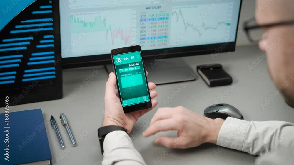 The guy in the office uses his bitcoin purse on the smartphone. In the frame of the hands on the phone, the man enters his electronic account and looks through the transactions.