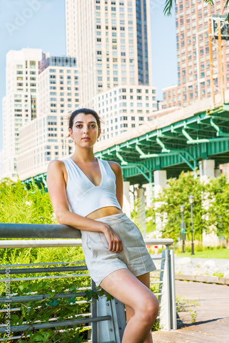 Woman Casual Summer Fashion in New York. Young pretty girl wearing white deep v neck crop tank top, shorts, standing by metal fence at street park, relaxing. High apartment buildings on background.