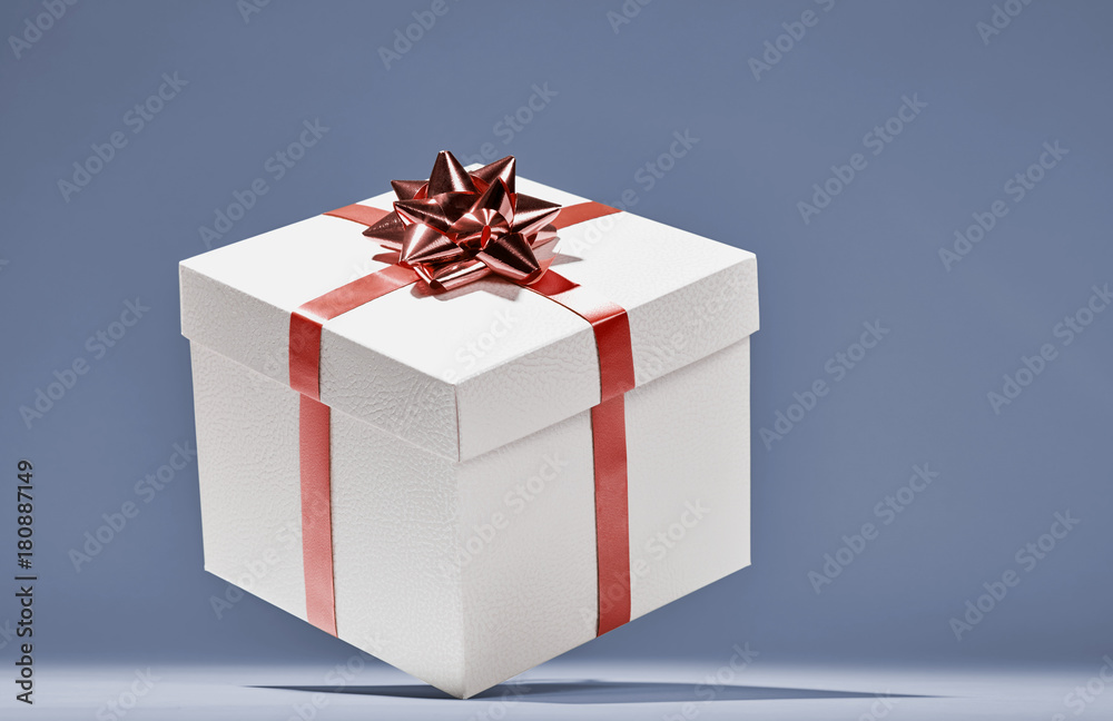Fototapeta premium White cube gift box with red stripes and bow floating in intimate blue background