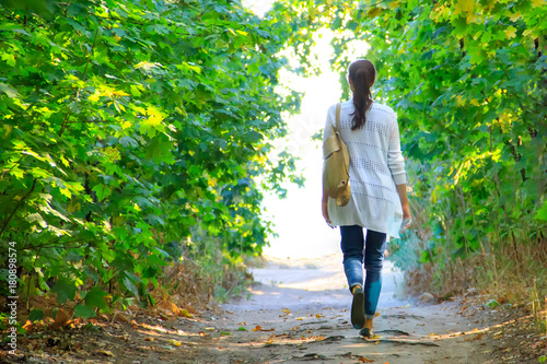 The girl walks along the path in the woods to the light in a white jacket and jeans.