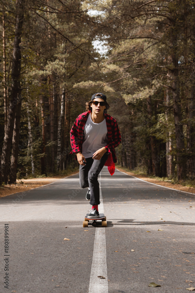 Obraz premium A young hipster in a cap and plaid shirt is riding his longboard on a country road in the forest
