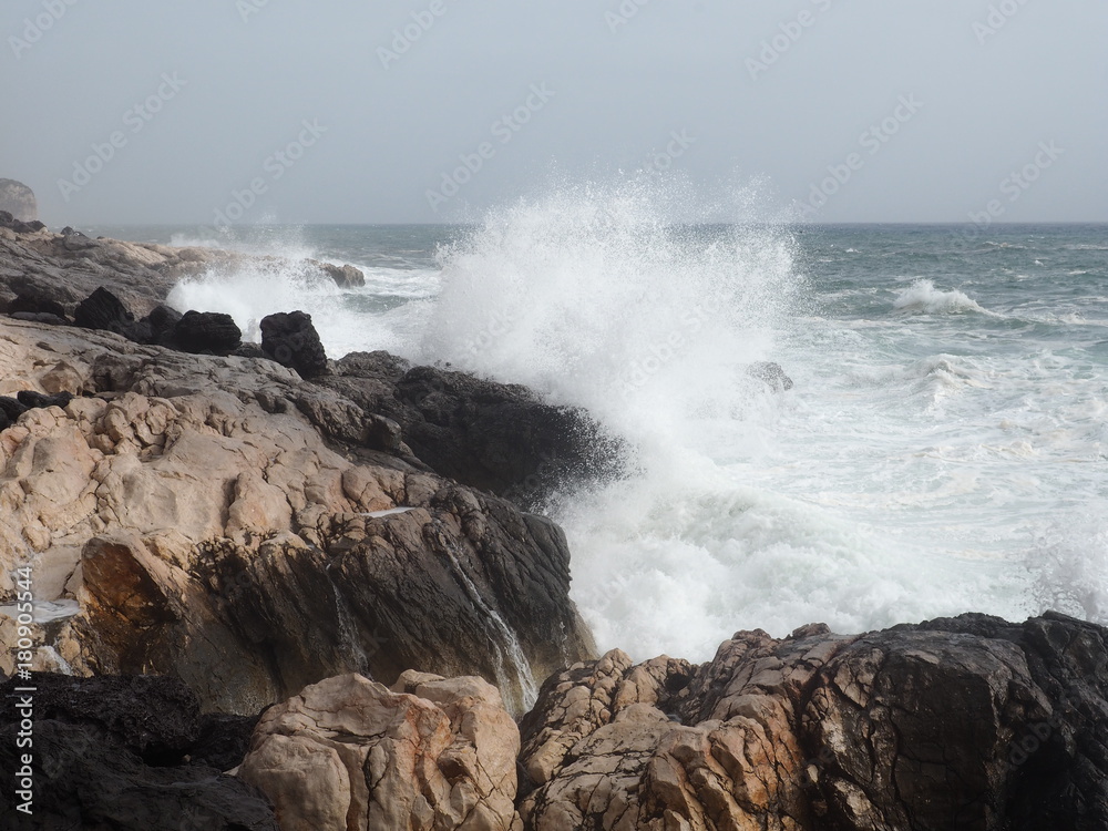 Eine schroffe Küste auf die eine große Welle trifft.