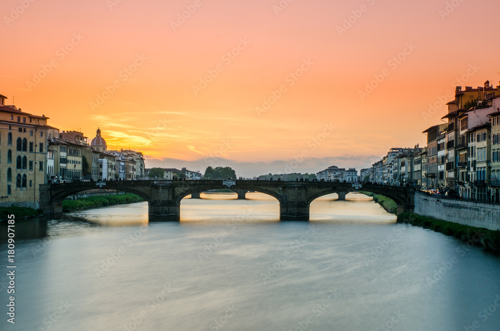 A long exposure of Ponte Santa Trinita and the River Arno, viewed from the Ponte Vecchio in Florence, Italy as the sun sets in the background lighting the sky up with stunning pink and orange hues.