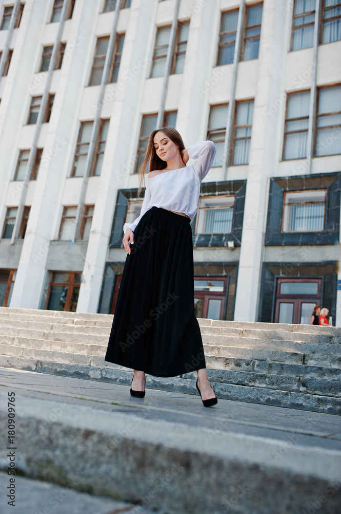 Portrait of a fabulous young successful woman in white blouse and broad black pants posing on the stairs with a huge white building on the background.