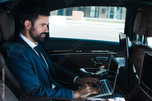 On the way to successful meeting. Side view of handsome young man using laptop with smile while sitting in car photo