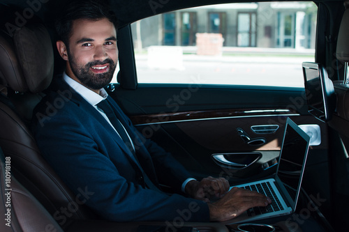 Leadership in his eyes. Side view of handsome young man using laptop and looking at camera with smile while sitting in car