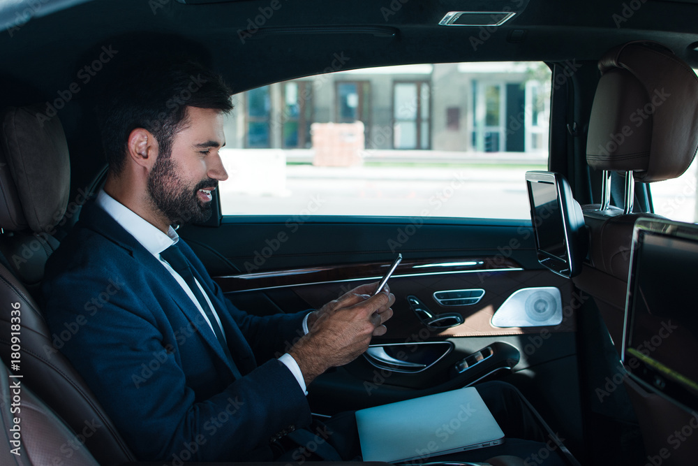 One more message. Side view of handsome young man using his smart phone with smile while sitting in car