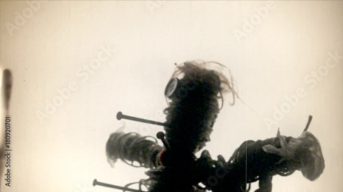Fake 8mm amateur film: the scay silhouette of a cursed voodoo doll, pierced by big rusty nails, over a white background.
 photo