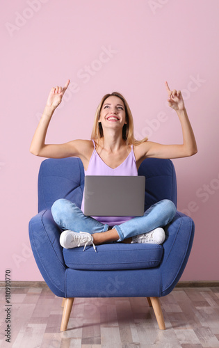 Young woman with laptop in armchair against pink wall