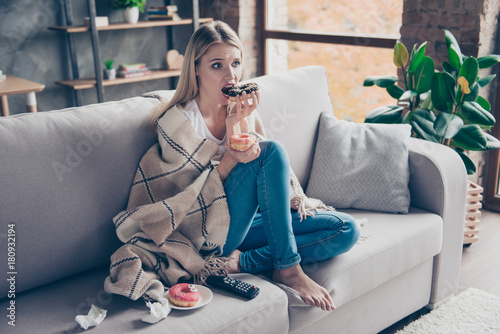 Beautiful blonde woman sitting on couch in living room under blanket eating chokolate donat watching something exciting interesting on television having health problem