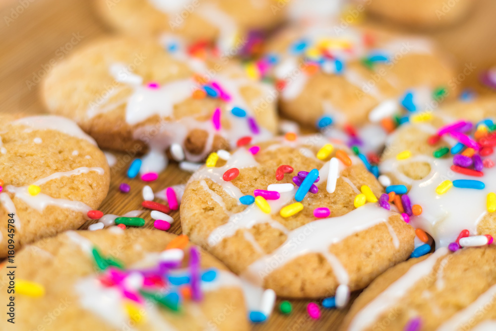 Freshly baked sugar cookies with white icing and rainbow colored sprinkles on wooden board, selective focus