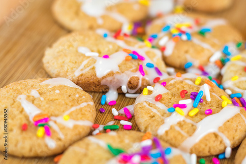 Freshly baked sugar cookies with white icing and rainbow colored sprinkles on wooden board, selective focus