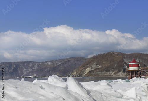 Ice hummocks on lake Baikal shore. Siberia winter landscape view with red and white lighthouse. Snow-covered ice of the lake. Big cracks in the ice floe. White clouds on blue sky