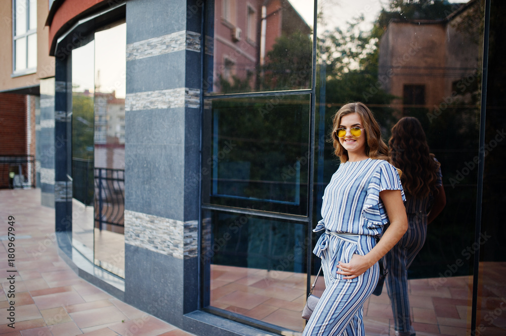 Portrait of a perfect young woman wearing striped overall and yellow sunglasses poses with her handbag on a balcony of a modern building in a town.