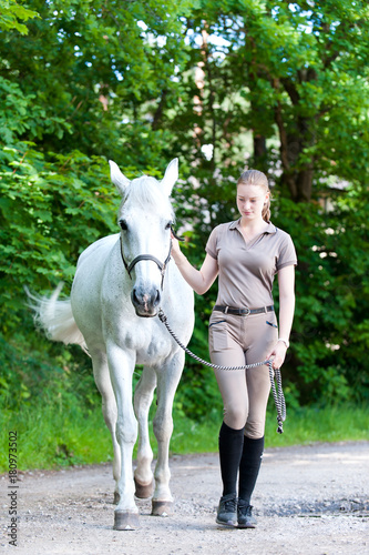 Young teenage blondy girl leading her favorite white horse.