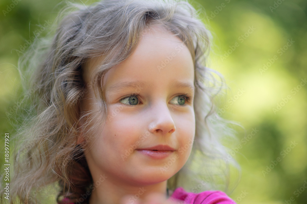 Beautiful little curly blonde girl, has happy fun cheerful smiling face, big blue eyes, long eyelashes. Portrait in green wood background Beautiful little curly blonde girl, has happy fun cheerful smiling face, big blue eyes, long eyelashes. Portrait in green wood background