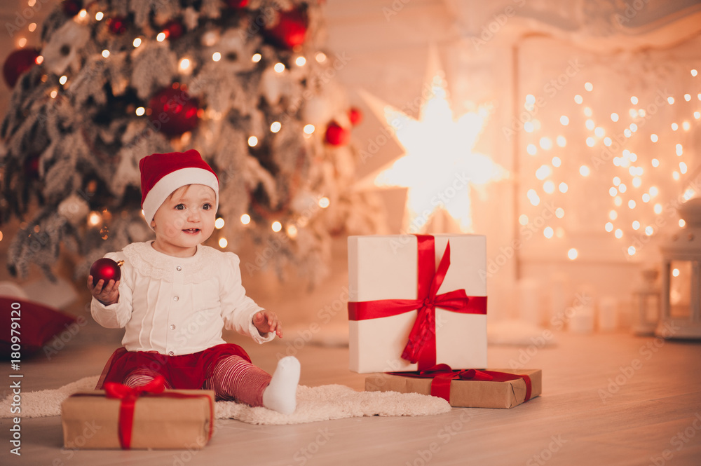 Cute baby holding Christmas ball wearing Santa Claus hat sitting with  presents over Christmas tree in room. Holiday season. Stock Photo | Adobe  Stock