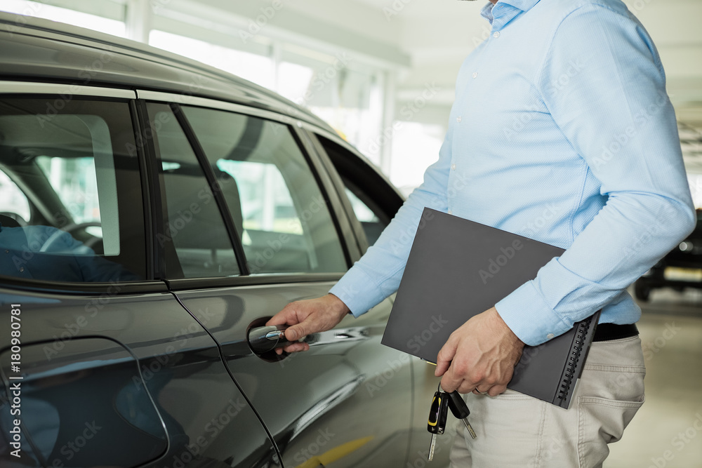 Close up photo of man opening a car door, holding a car door handle with one hand and folder and car keys in other hand