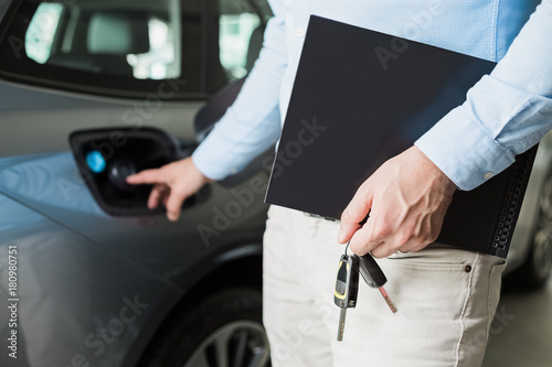 Close up of male adult car dealer standing indoors at car dealership,holding a folder and car keys in his hand, poiting finger towards a fill spout, talking about gas consumption