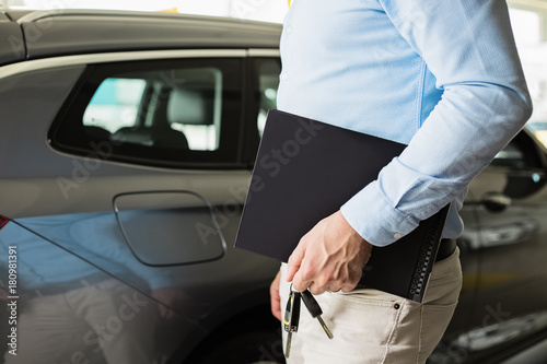 Close up of male adult car dealer standing indoorts at car dealership,holding a folder and car keys in his hand with a new car behind him © Zdenka