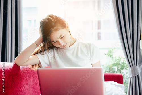Asian teenage girl using a laptop computer to check her orders online and drinking coffee on a red sofa in her home. She stressed that the sales slump.