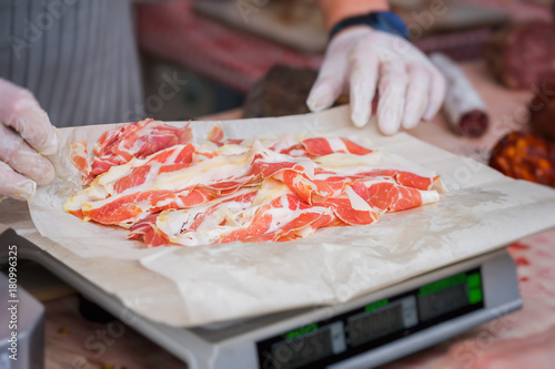 Close-up of thin slices of Italian ham, prosciutto crudo weight on scale, grocery store, hands of seller. Selective focus