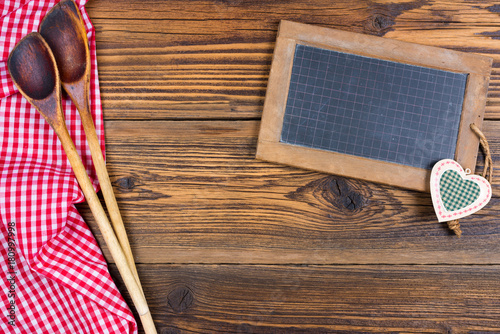 Zwei alte Holzlöffel und eine alte Schiefertafel sowie ein rot weiß kariertes Stofftuch liegen auf einem rustikalen Holz Hintergund mit Textfreiraum zum selber gestalten